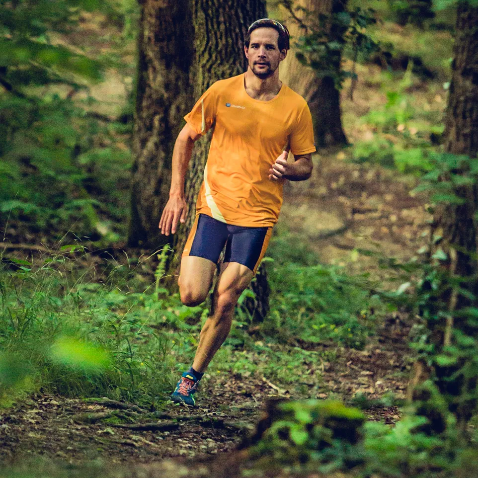 Un uomo corre su un sentiero boscoso indossando una maglia da corsa arancione e pantaloncini da corsa blu scuro.