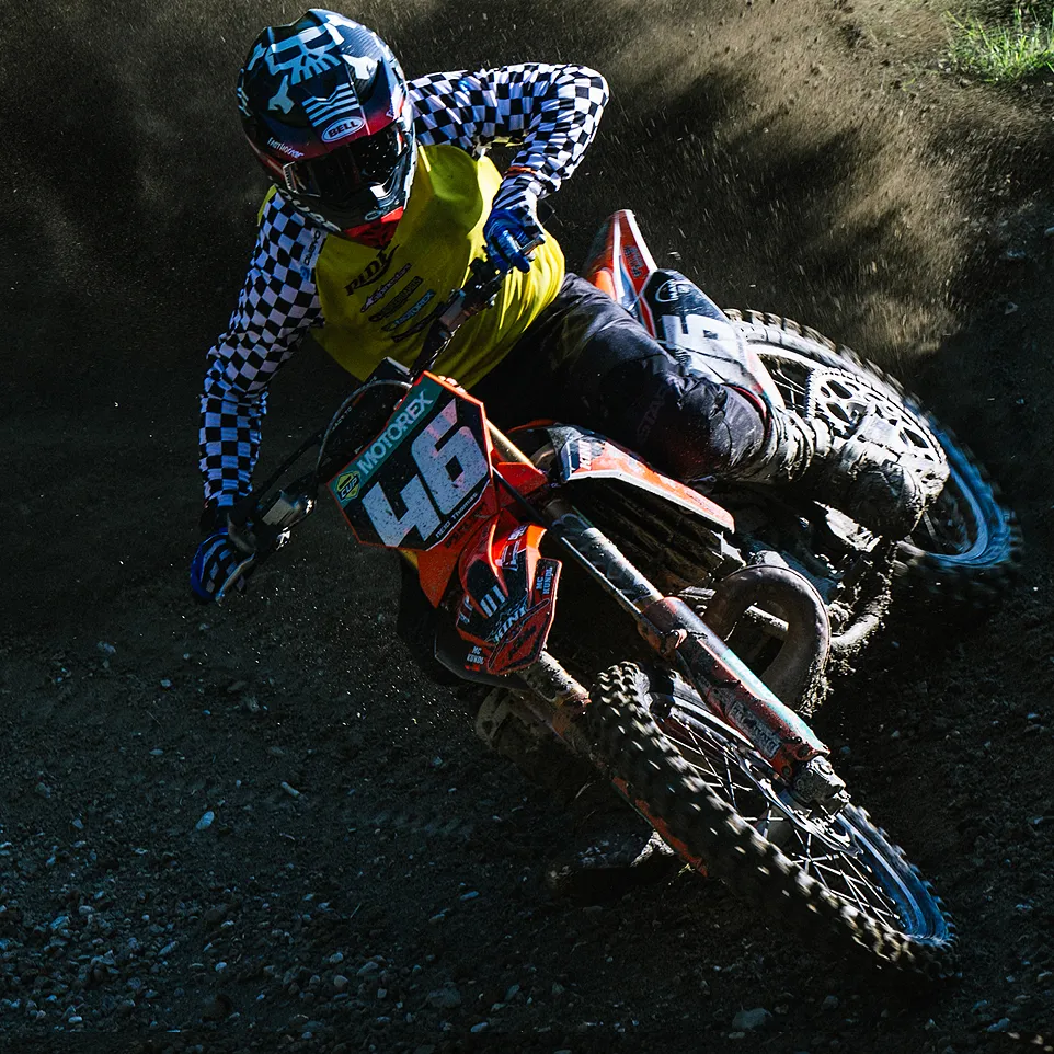 Un pilota di motocross in una giacca gialla con maniche a scacchi neri e bianchi e un casco guida una moto arancione su un circuito polveroso.